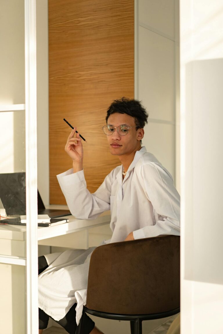 A man in a white robe sits at a desk using a laptop in a sunlit room, looking thoughtfully.